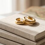 Close-up macro photograph of a pair of chunky, polished gold hoop earrings resting atop a stack of linen-bound travel journals in bright daylight.