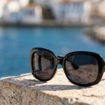 Close-up shot of oversized black square sunglasses resting on a weathered stone ledge, with a blurry background of a Mediterranean village and turquoise sea.