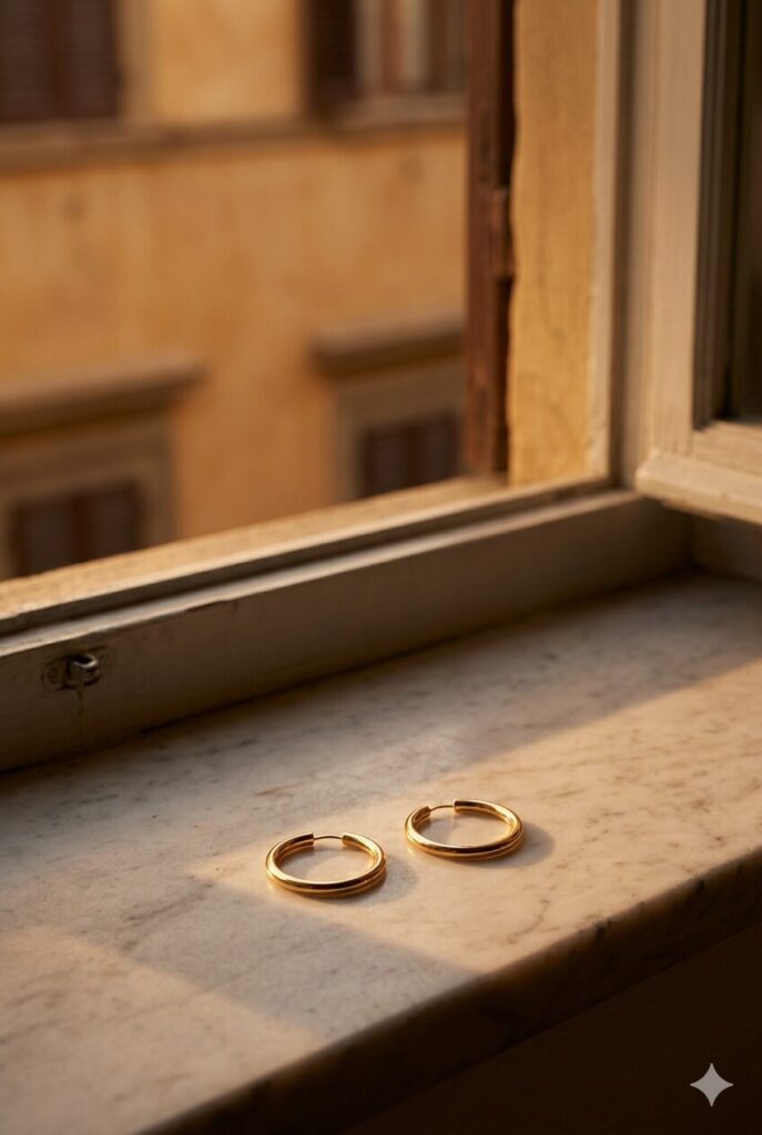 Pair of symmetrical gold hoop earrings resting on a worn marble windowsill in Florence during the golden hour, overlooking ochre buildings.