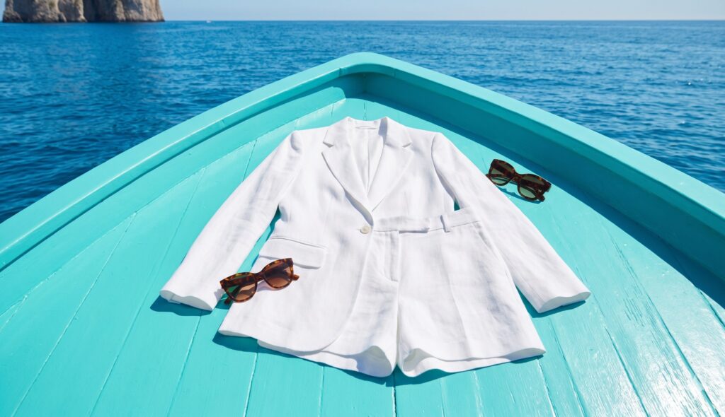 Caprese white women's linen blazer and shorts suit laid flat on turquoise Capri boat deck with tortoiseshell sunglasses and azure Tyrrhenian Sea in the background: Campania travel capsule wardrobe colour palette