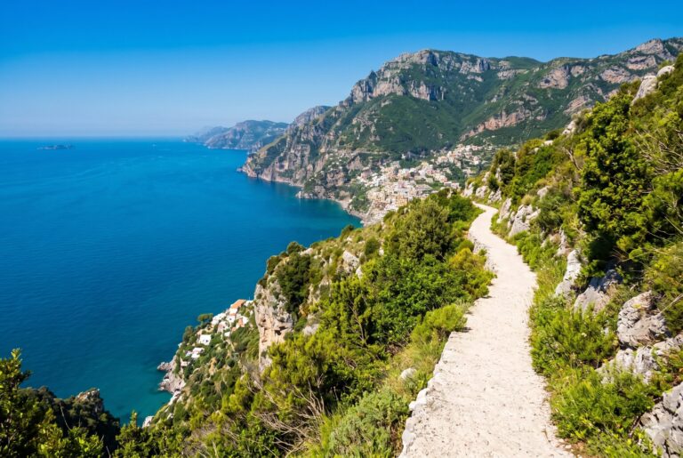 The Path of the Gods trail running along the dramatic cliff edge ridge above the Amalfi Coast with vivid green Mediterranean scrub either side and the deep aquamarine Tyrrhenian Sea Positano and Capri visible far below under a cobalt blue sky