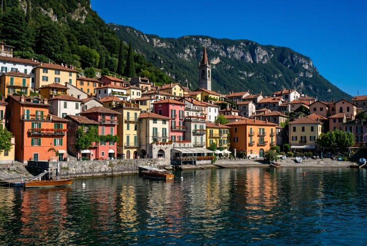 The colourful Varenna waterfront on Lake Como seen from the water with vivid terracotta orange pale yellow and deep rose pink building facades with green shutters and red geraniums rising directly from the deep blue-green lake water and steep dark green forested mountains behind