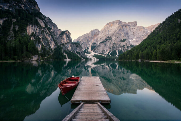 Lago di Braies in the Dolomites at early dawn with a single red wooden rowboat at the dock the pale dolomite peaks reflected in the deep emerald green lake water and pre-dawn indigo and gold sky above