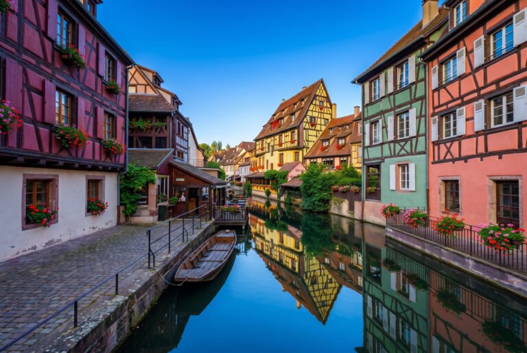 The Petite Venise canal quarter in Colmar Alsace in early morning light with vivid deep burgundy red forest green mustard yellow and coral pink half-timbered facades reflecting in the still canal water and window boxes of red geraniums above the stone quayside