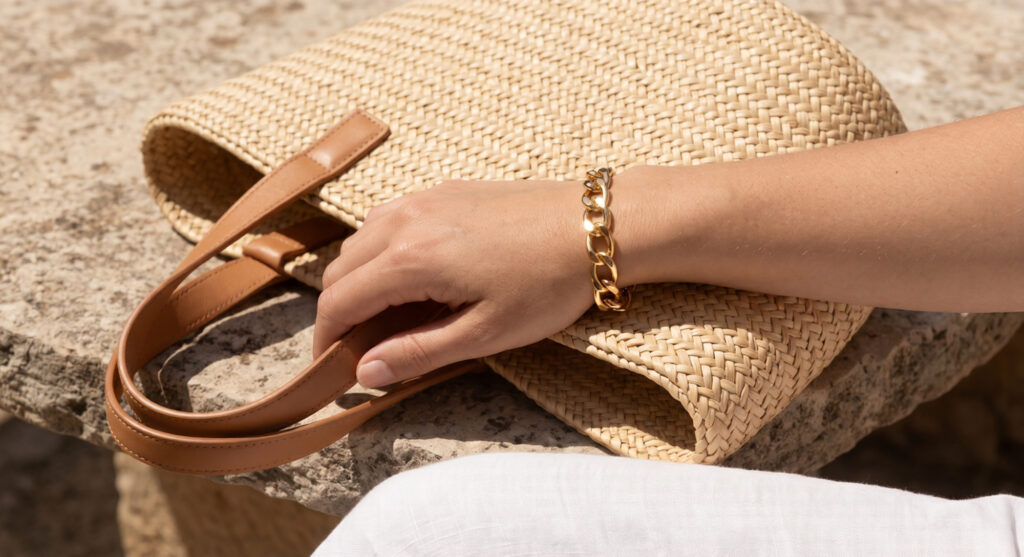 A close-up of a person wearing a gold link bracelet, resting an arm on a natural woven straw and raffia tote bag outdoors on a sunny day.