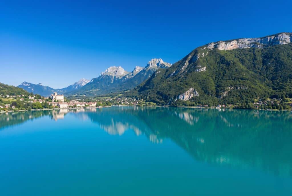 Aerial view of Lake Annecy in the French Alps with extraordinary turquoise-blue lake water reflecting the steep forested Aravis and Bauges mountain slopes on the far shore and the medieval old town of Annecy with the ch&acirc;teau on the hillside visible at the northern end under a cobalt blue sky