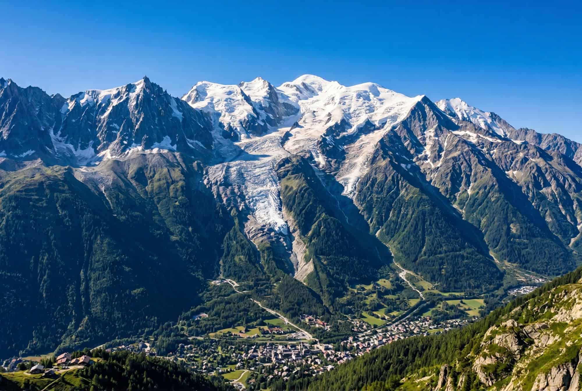 The Mont Blanc massif from above the Chamonix valley in the French Alps with the pale grey-white glacier and ice cap at 4808 metres the dramatic Aiguilles rock spires the Mer de Glace glacier and the deep green Chamonix valley far below under a deep cobalt blue sky