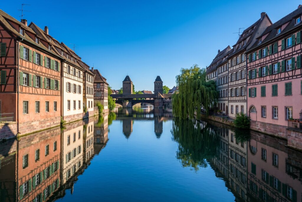 The Petite France quarter in Strasbourg with the Ponts Couverts covered bridges and medieval towers in the background and tall half-timbered tanners' houses in faded terracotta pale cream and soft rose with dark timber frames and green shutters reflected in the still Ill river water