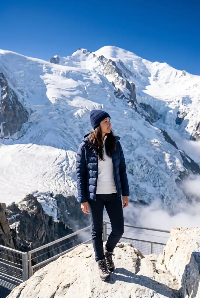 Stylish woman in navy technical puffer jacket and white merino base layer standing on the Aiguille du Midi viewing platform at 3842 metres with the full pale grey-white Mont Blanc glacier and ice cap rising directly behind her against a deep cobalt blue sky