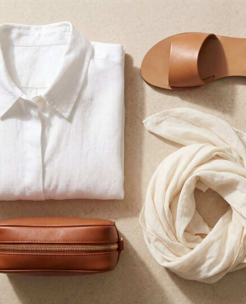 Close-up flat lay of white linen shirt, leather sandal, cognac crossbody bag and ivory scarf on pale stone