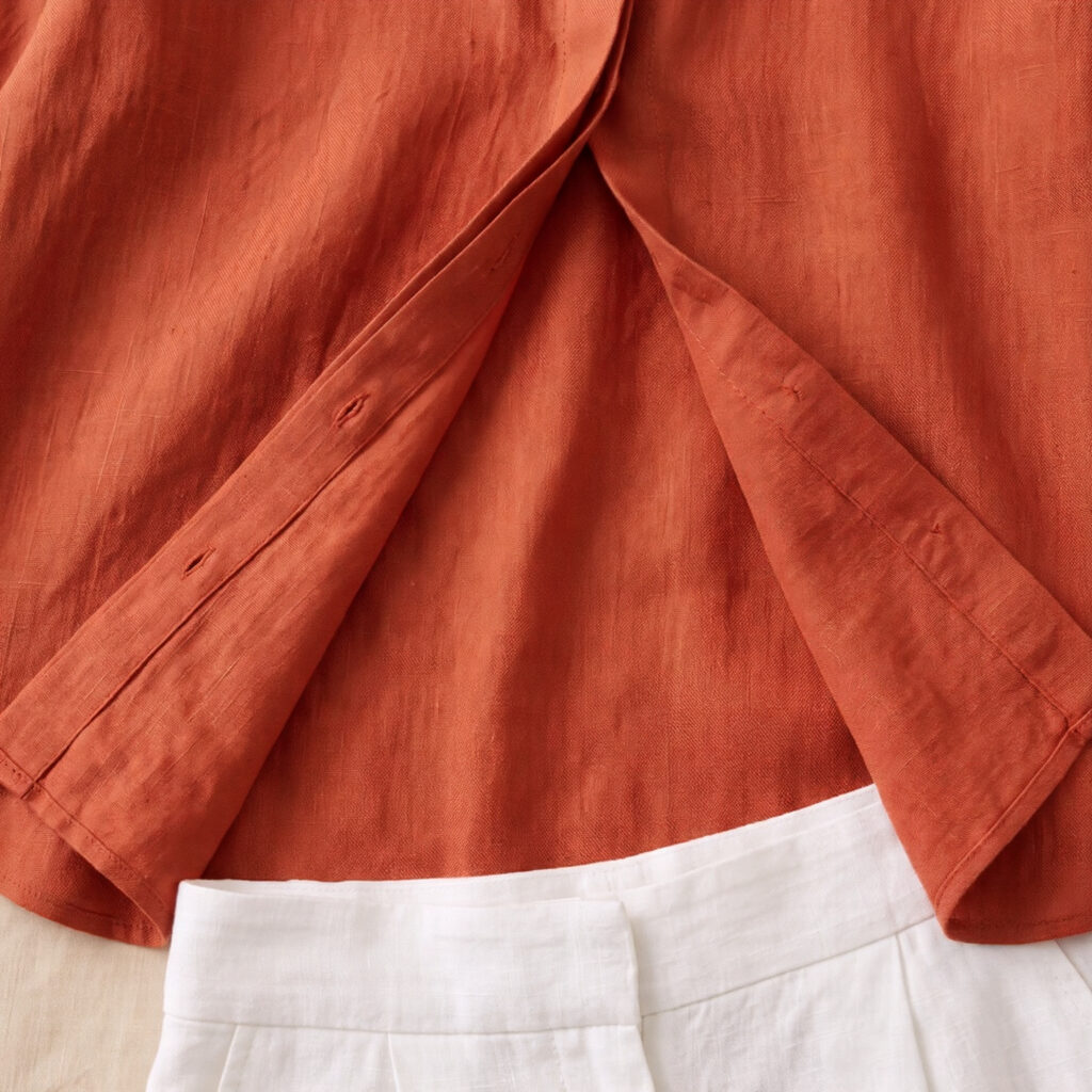 Close-up of terracotta oversized linen shirt hem and open placket with bleached white linen trouser waistband visible below