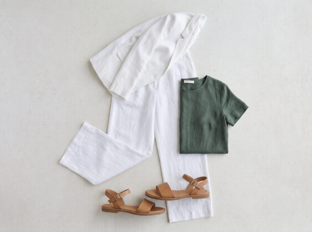 Flat lay of Costa Brava coastal path capsule outfit: bleached white wide-leg linen trousers, deep pine green linen top, flat tan ankle-strap sandals, and white linen relaxed blazer on limestone surface