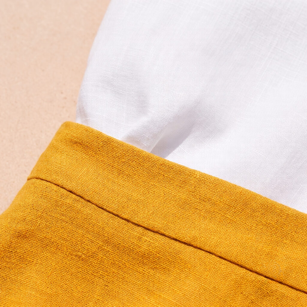Close-up of deep saffron yellow linen midi skirt waistband meeting white linen shirt tucked inside, vivid colour contrast and linen weave detail in strong natural light