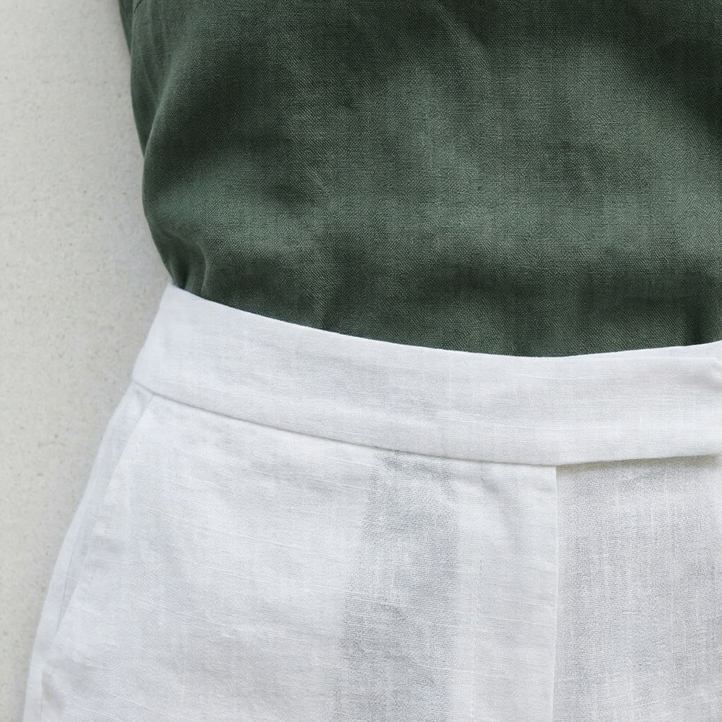 Close-up of bleached white linen trouser waistband meeting deep pine green linen top tucked inside, linen weave and colour contrast in coastal daylight