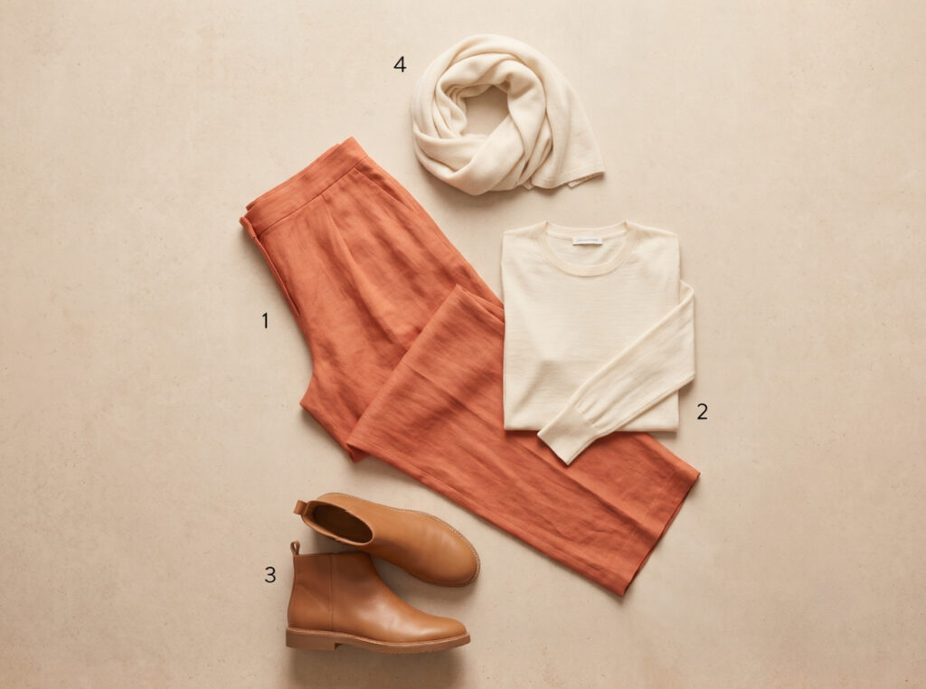 Flat lay of Granada medina and evening capsule outfit: Darro terracotta wide-leg linen trousers, ivory merino long-sleeve top, flat tan ankle boots, and ivory wool wrap on pale stone surface