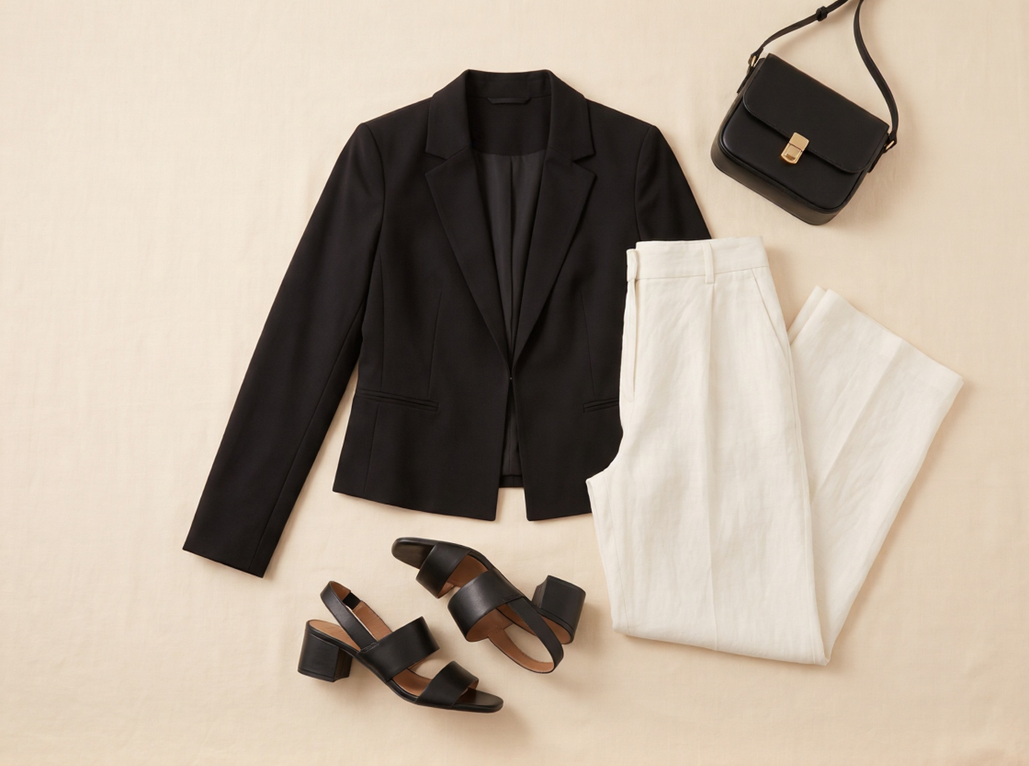 Flat lay of Madrid evening capsule outfit: fitted black blazer, ivory wide-leg linen trousers, black block-heel sandals, and black leather crossbody bag on cream linen surface