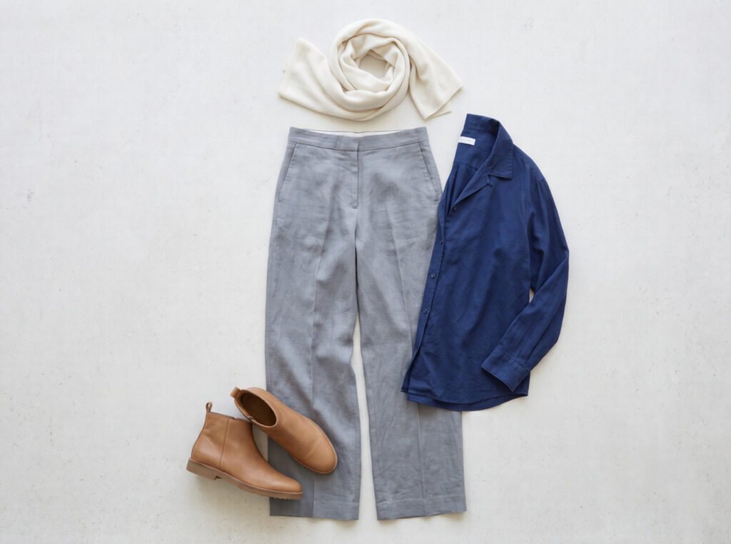 Flat lay of Granada Albaicin capsule outfit: sierra grey wide-leg linen trousers, indigo cotton shirt, flat tan leather ankle boots, and ivory cashmere wrap on pale stone surface