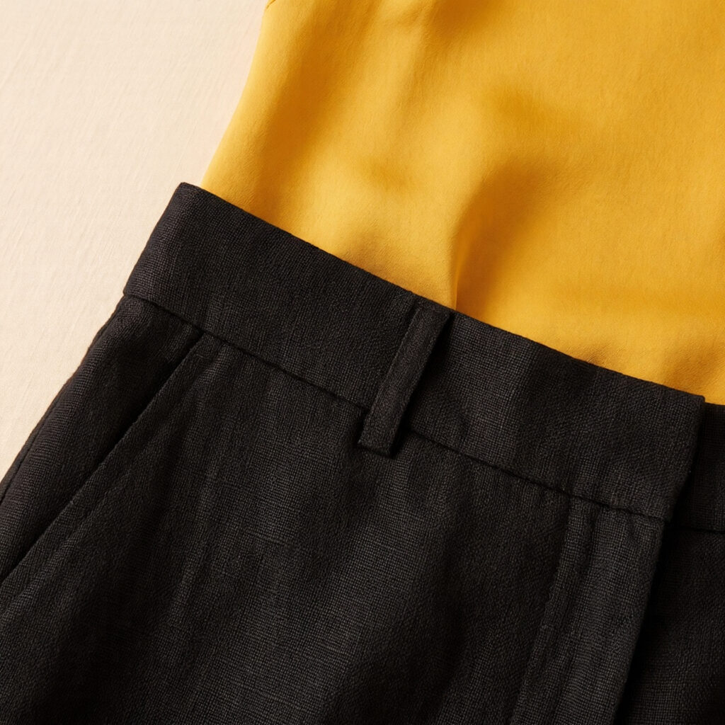 Close-up of black mid-weight linen wide-leg trouser waistband meeting saffron yellow silk camisole tucked inside, vivid colour and fabric contrast in warm evening light